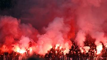 Tłum kibic&oacute;w na stadionie odpalający race i świece dymne, tworząc czerwone obłoki dymu.