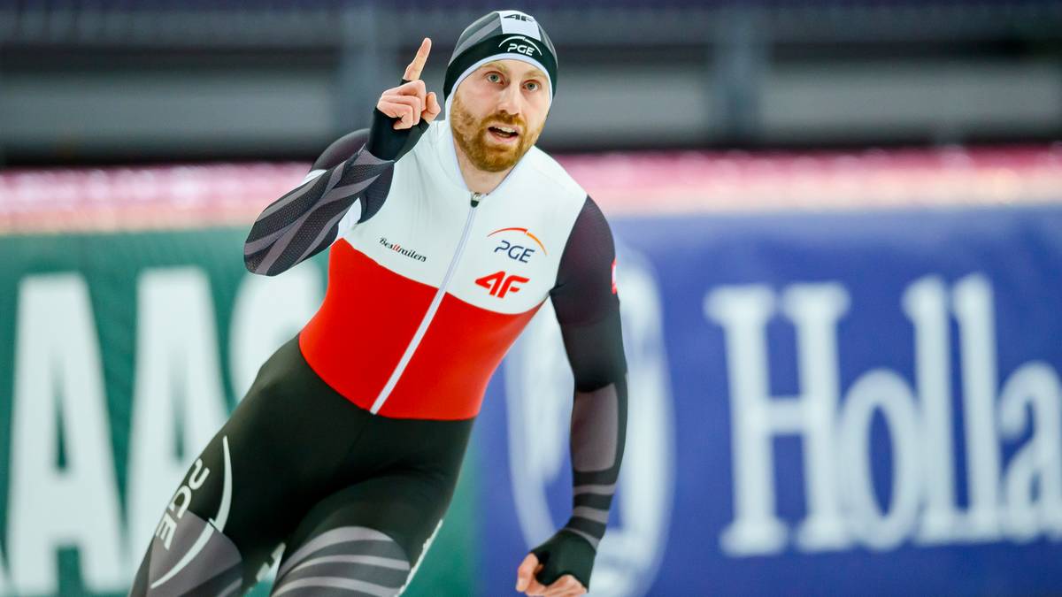 Łyżwiarz szybki z brodą i czapką na głowie, w stroju sportowym z elementami czerwonymi i białymi, wskazuje palcem w górę.