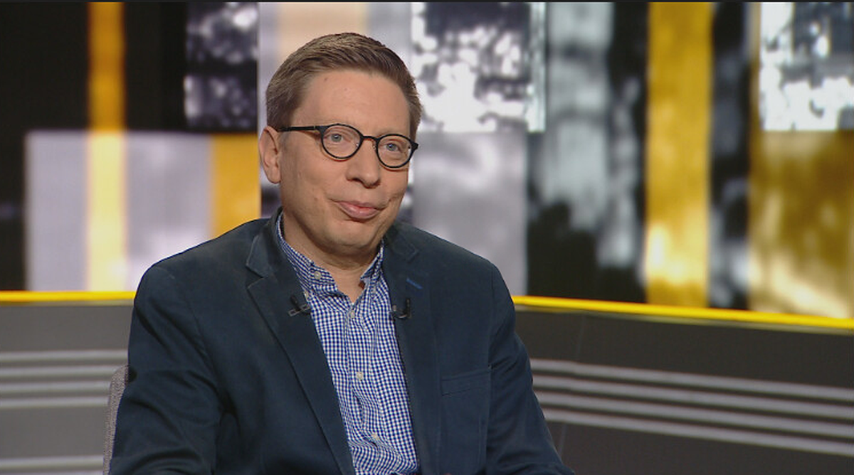 Mężczyzna w okularach siedzi w studiu telewizyjnym, rozmawiając.