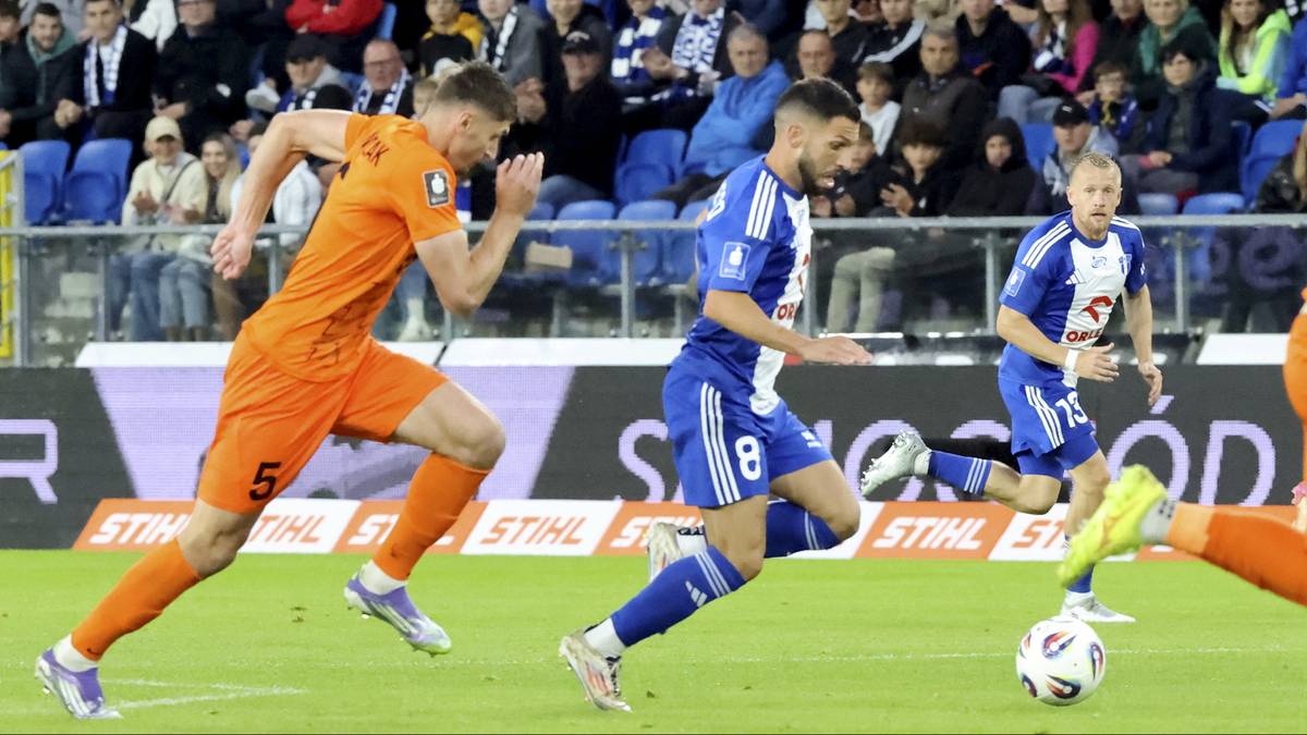 Dw&oacute;ch piłkarzy w trakcie meczu, jeden w stroju pomarańczowym, drugi w niebiesko-białym, biegnie w kierunku piłki na zielonej murawie.