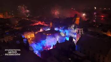 Widok z lotu ptaka na miasto nocą, z iluminacjami świetlnymi na budynkach i pokazem fajerwerk&oacute;w na niebie.