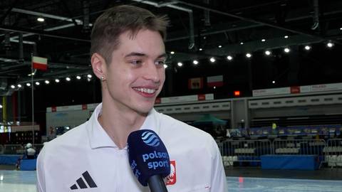 Młody mężczyzna z jasnymi włosami uśmiecha się, trzymając mikrofon Polsat Sport.