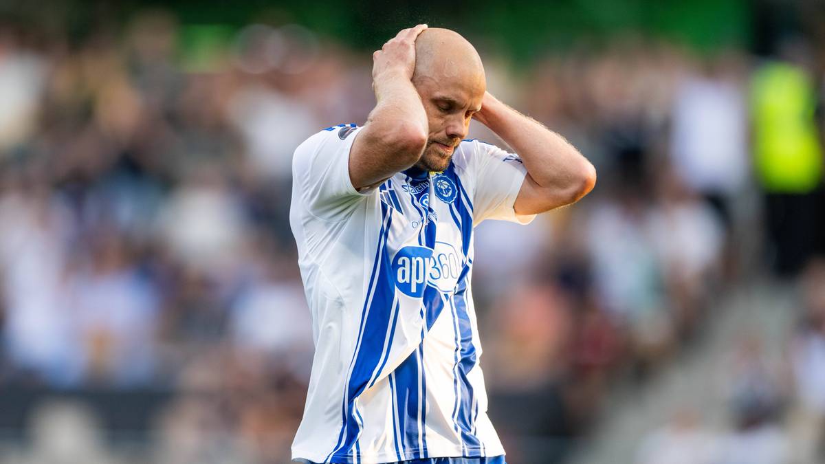 Łysy piłkarz w białej koszulce z niebieskimi pasami trzyma się za głowę na tle stadionu.