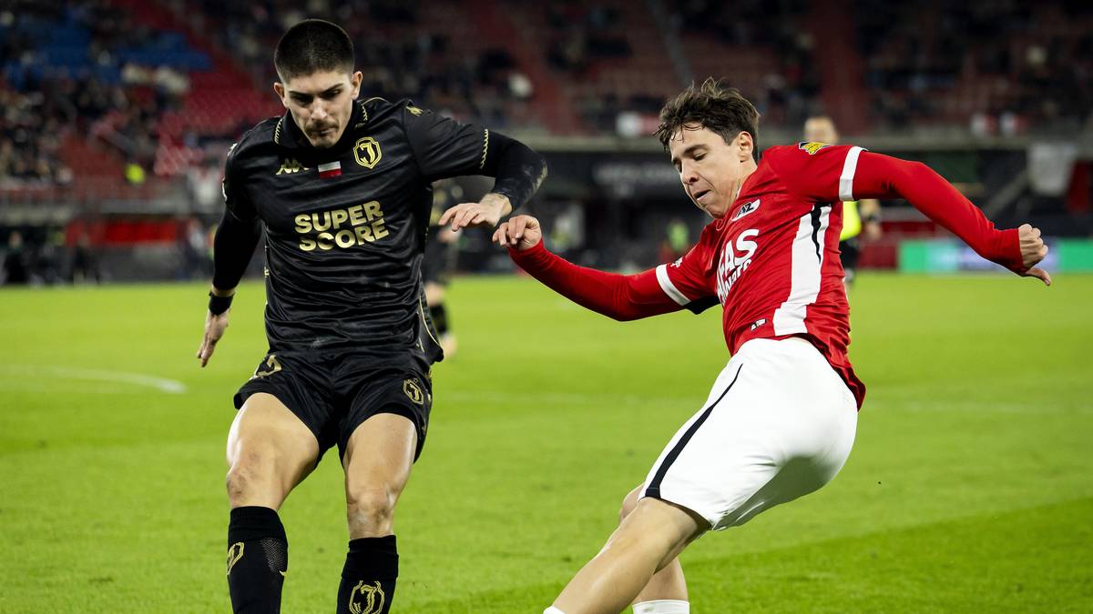 Dw&oacute;ch piłkarzy w akcji na boisku, jeden w czarnej koszulce z ż&oacute;łtymi elementami, drugi w czerwonej koszulce z białymi spodenkami.