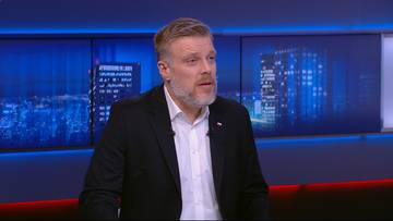 Mężczyzna w średnim wieku z brodą, ubrany w czarną marynarkę i białą koszulę, mówi patrząc w kamerę. Tło stanowią ekrany z widokiem nocnego miasta.