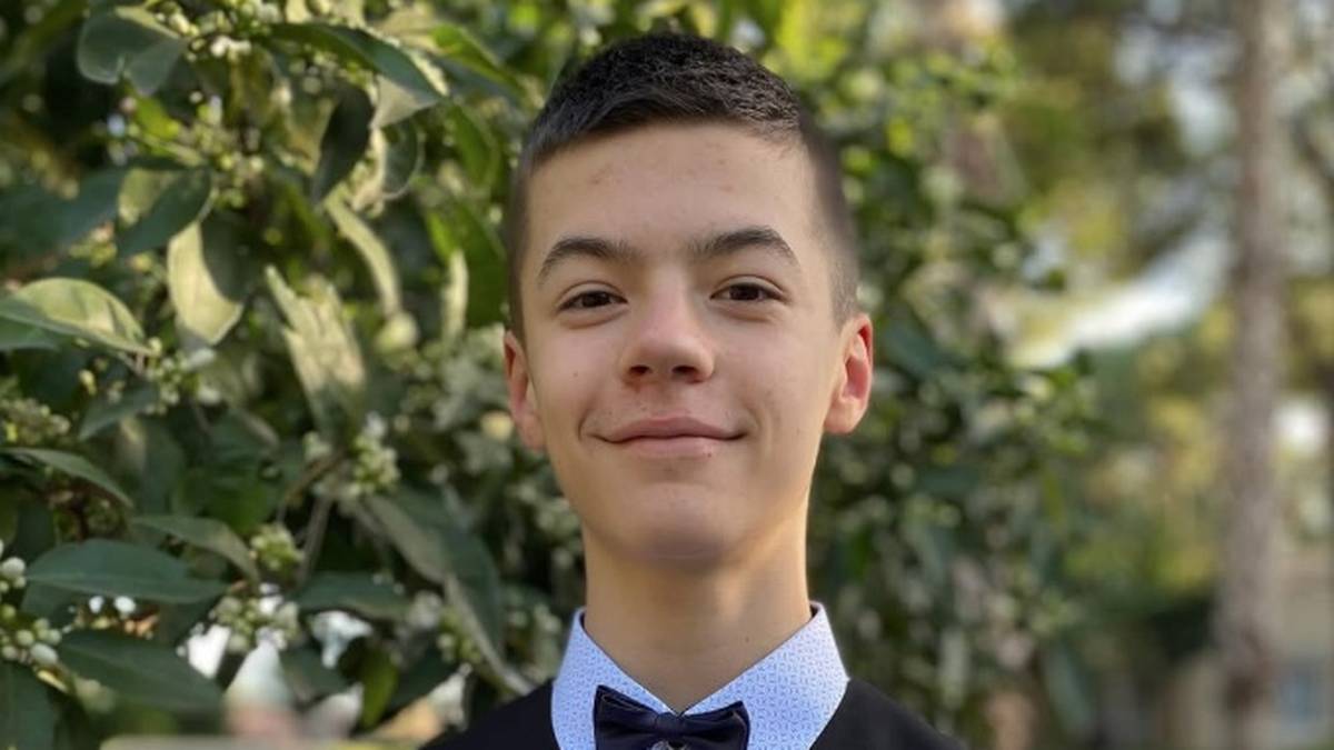 Portret uśmiechniętego 14-letniego chłopca w kamizelce i muszce na tle zielonych liści.