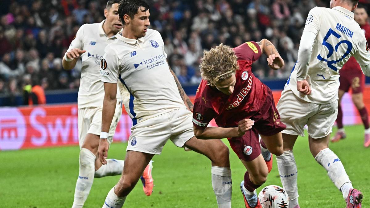 Trzech piłkarzy w akcji podczas meczu piłki nożnej. Dwóch zawodników w białych strojach walczy o piłkę z graczem w czerwonym stroju. W tle widać trybuny stadionu.