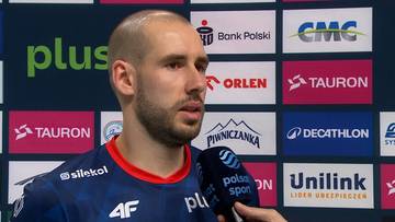 Mężczyzna z krótką brodą w koszulce sportowej, udzielający wywiadu na tle sponsorów.