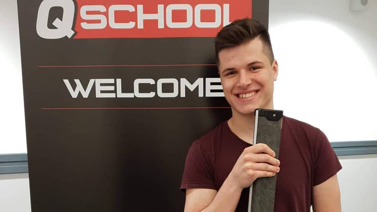 Uśmiechnięty młody mężczyzna stoi przed tablicą z napisem "Q SCHOOL WELCOME" i trzyma przedmiot przypominający certyfikat lub nagrodę.