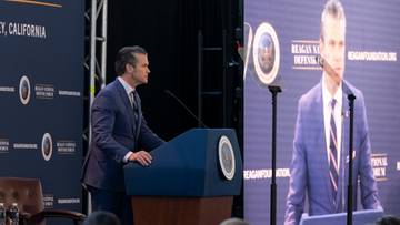 Pete Hegseth przemawia za mównicą, podczas gdy na dużym ekranie wyświetlany jest jego portret.