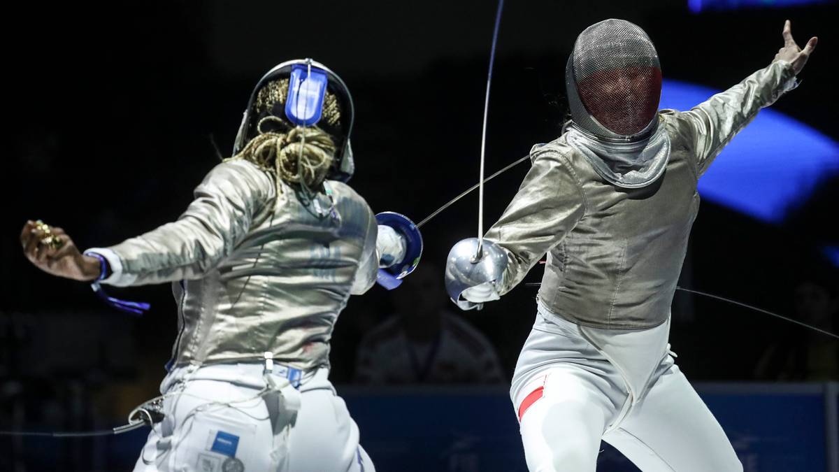 Dwie zawodniczki szermierki w białych strojach i maskach na twarzach rywalizują podczas turnieju. Jedna z nich ma wyciągniętą prawą rękę z medalem.