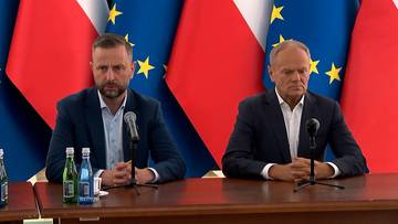 Dw&oacute;ch mężczyzn w garniturach siedzi przy stole konferencyjnym. Na ścianie za nimi widnieją flagi Polski i Unii Europejskiej.