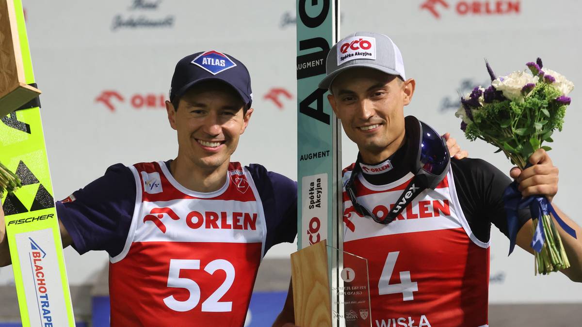 Dwóch uśmiechniętych polskich skoczków narciarskich stoi na podium, trzymając narty i bukiet kwiatów.