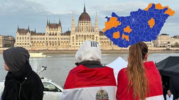 Trzy osoby z widokiem od tyłu, ubrane w narodowe barwy Węgier, stoją na tle Parlamento Węgierskiego w Budapeszcie.