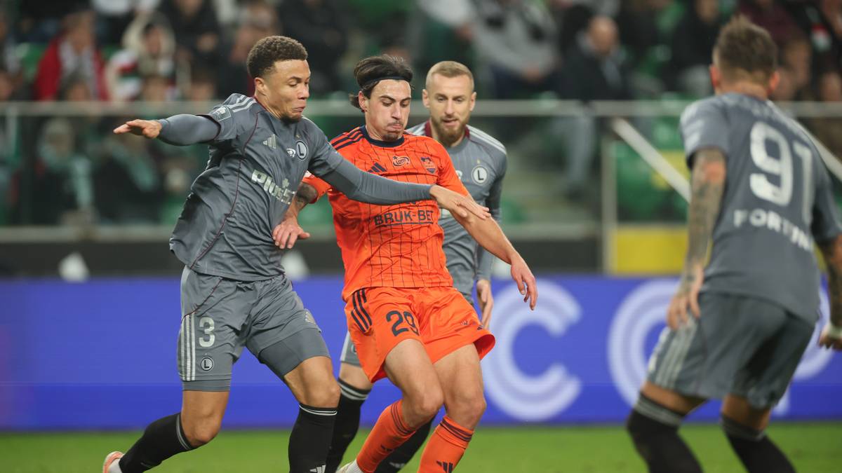 Trzech piłkarzy na boisku podczas meczu, dw&oacute;ch w szarych strojach i jeden w pomarańczowym, walczących o piłkę.