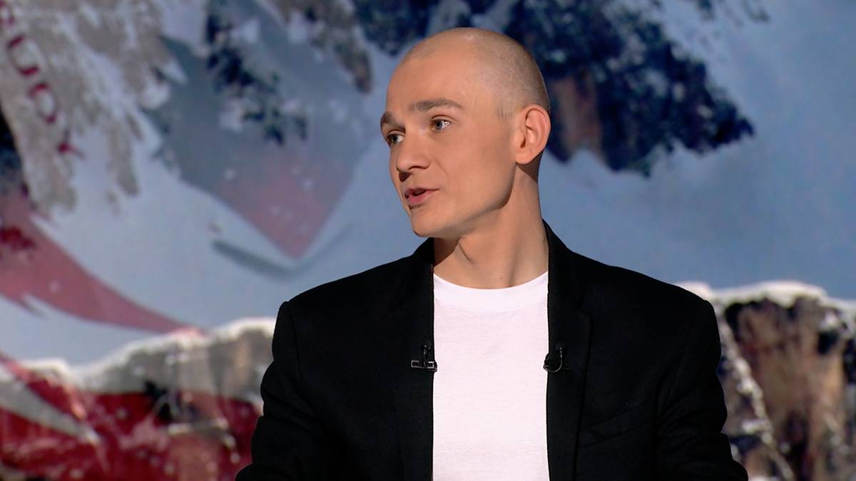 Portret mężczyzny z łysą głową w czarnej marynarce i białej koszulce, patrzącego w bok, na tle zimowego krajobrazu z g&oacute;rami i śniegiem.