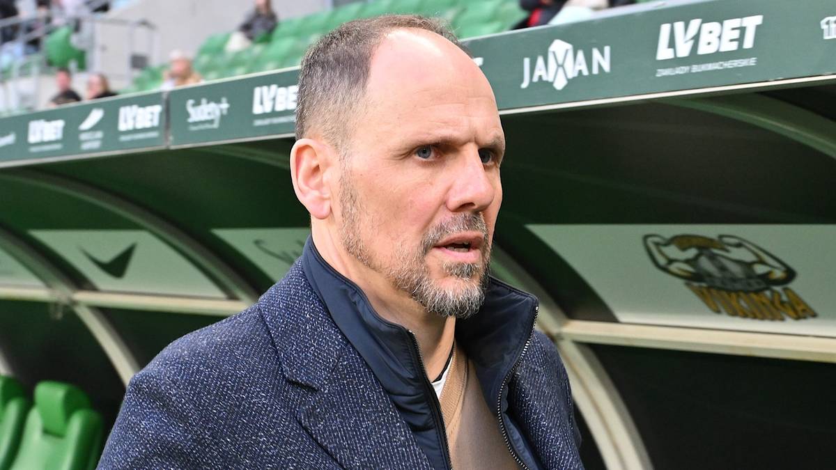 Trener GKS Tychy w garniturze stoi na tle zielonych siedzeń ławki rezerwowych.