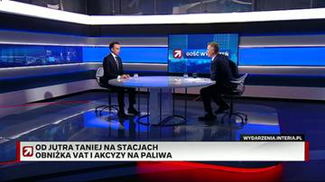 Dw&oacute;ch mężczyzn w garniturach siedzi przy stole w studiu telewizyjnym, podczas gdy na ekranie wyświetlany jest komunikat o obniżce cen paliw.