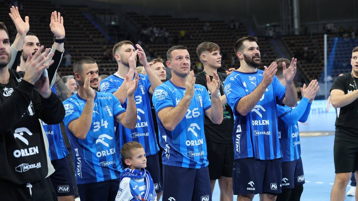 Grupa mężczyzn w niebieskich strojach sportowych, niekt&oacute;rzy z numerami na koszulkach, klaszcze i patrzy w g&oacute;rę. W tle widać trybuny z widzami. Na pierwszym planie stoi chłopiec w niebieskim stroju.