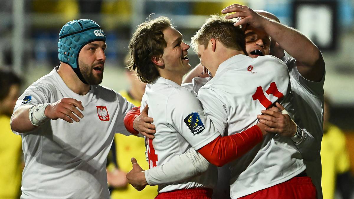 Czterech graczy rugby w białych strojach z polskim godłem świętuje zwycięstwo, obejmując się i śmiejąc. Jeden z zawodników ma na głowie niebieski ochraniacz.