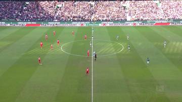 Boisko piłkarskie z zawodnikami obu drużyn, jeden zesp&oacute;ł w czerwonych strojach, drugi w zielonych. W tle trybuny wypełnione widzami.