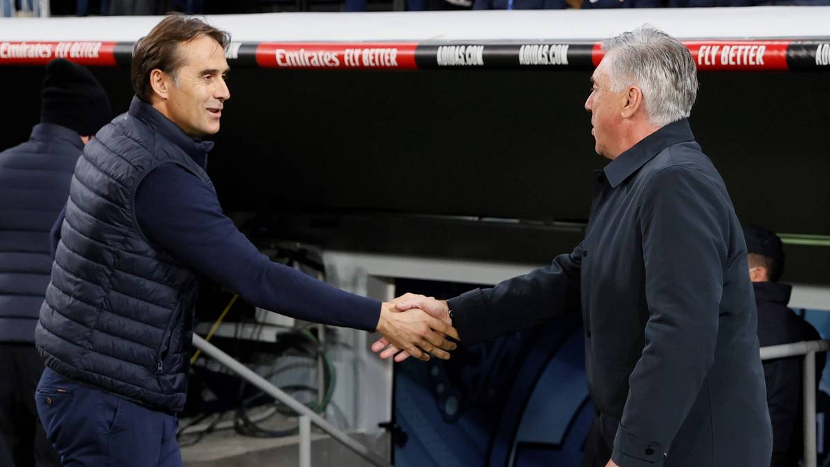 Dwóch mężczyzn w strojach sportowych, prawdopodobnie trenerzy piłkarscy, ściska sobie dłonie na tle trybun stadionu.