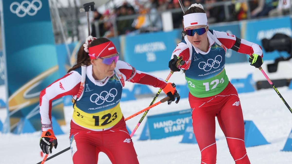 Dwie kobiety w strojach biatlonowych biegnące po śniegu, z karabinami na plecach i kijami w dłoniach. Na strojach widoczne są numery startowe i logo igrzysk olimpijskich.