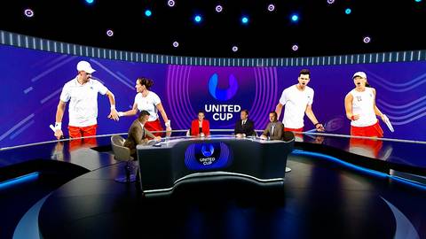 Zdjęcie przedstawia scenę z turnieju tenisowego United Cup, z zawodnikami na ekranach i komentatorami przy stole.