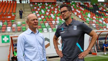 Dw&oacute;ch mężczyzn w strojach sportowych stoi na boisku piłkarskim przed trybunami.