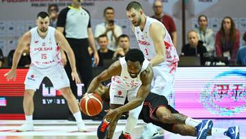 Dw&oacute;ch zawodnik&oacute;w drużyny Polski w białych strojach walczy o piłkę podczas meczu koszyk&oacute;wki. Jeden z nich klęczy na parkiecie, a drugi pr&oacute;buje odebrać mu piłkę, z pomocą trzeciego zawodnika pr&oacute;bującego podnieść klęczącego.