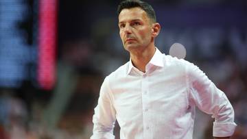 Mężczyzna w białej koszuli, prawdopodobnie trener sportowy, spogląda w bok z poważną miną.