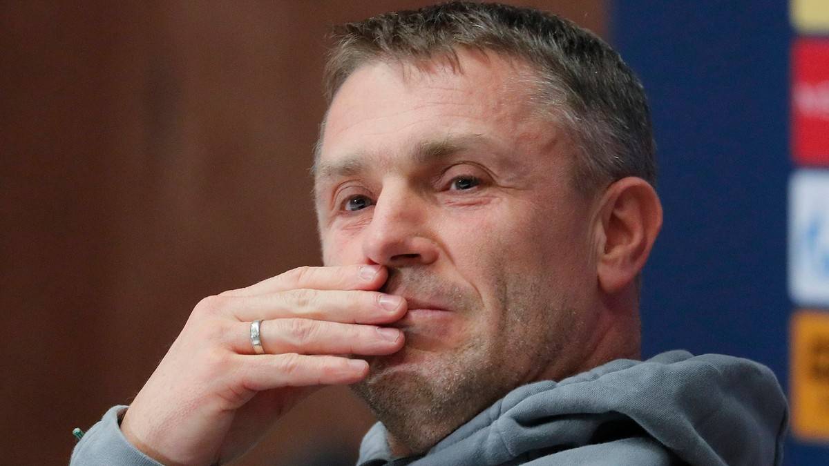 Mężczyzna w szarym swetrze, zakrywający usta dłonią.