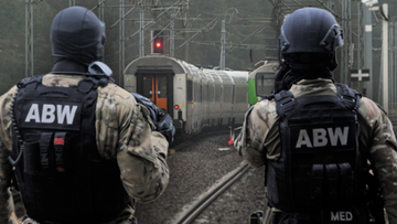 Dwóch funkcjonariuszy ABW w kamizelkach taktycznych z napisem ABW stoi tyłem do widza i obserwuje tory kolejowe, na których stoi pociąg.