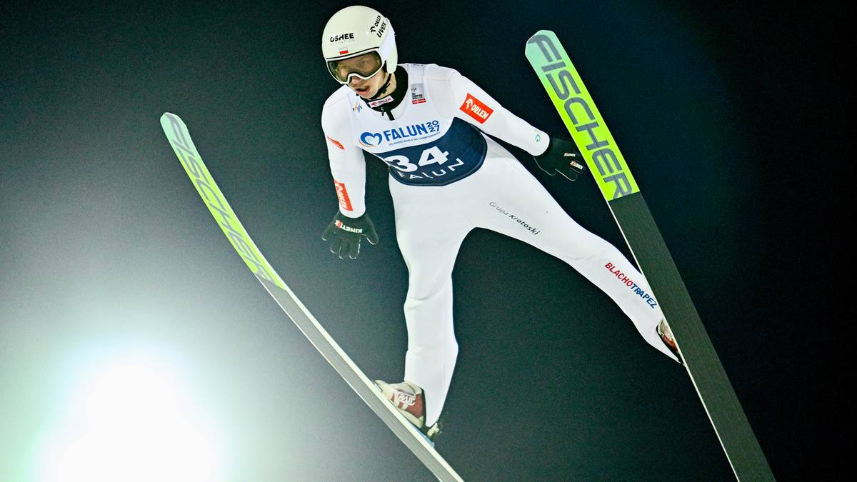 Skoczek narciarski w locie, ubrany w biały kombinezon z numerem 34, na tle ciemnego nieba.