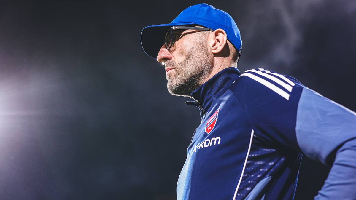 Mężczyzna w niebieskiej czapce i okularach przeciwsłonecznych, ubrany w dres sportowy, patrzy w bok z determinacją. W tle widać światła stadionu.