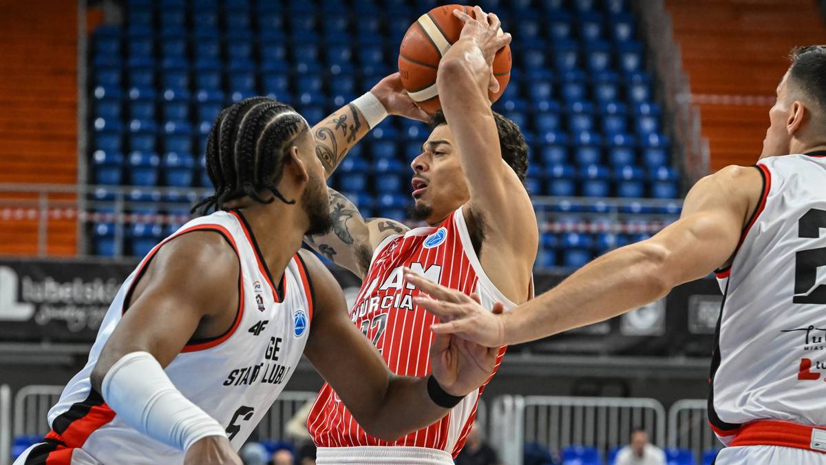 Koszykarz w czerwono-białej koszulce próbuje rzucić piłką do kosza, otoczony przez dwóch przeciwników w białych koszulkach.