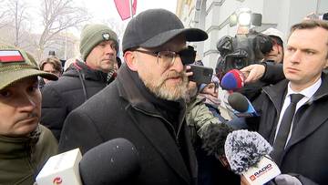 Grzegorz Braun w czapce i okularach przemawia do grupy dziennikarzy trzymających mikrofony.