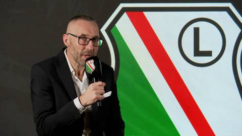 Marek Papszun w okularach i czarnym garniturze, trzymający mikrofon, stoi na tle logo Legii Warszawa.