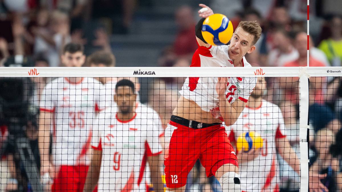 Siatkarz w biało-czerwonej koszulce z numerem 12 wykonuje zagrywkę, piłka w powietrzu, w tle inni zawodnicy i trybuny.