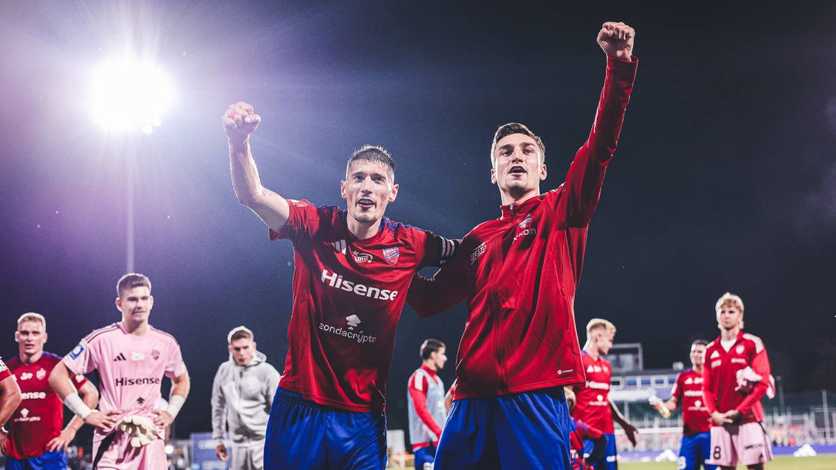 Dwóch piłkarzy w czerwonych strojach świętuje zwycięstwo, unosząc ręce w górę i obejmując się. W tle widać innych zawodników i sztuczne światła stadionu.