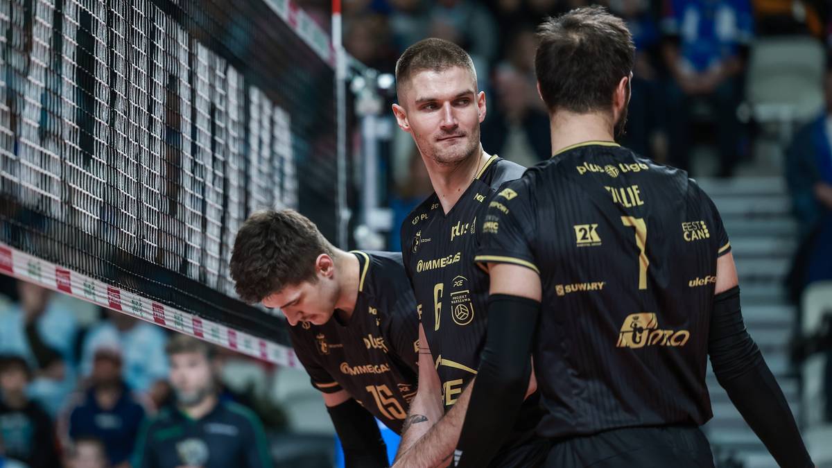 Trzech siatkarzy w czarnych strojach z ż&oacute;łtymi elementami rozmawia przy siatce podczas meczu.