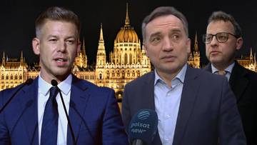 Dw&oacute;ch mężczyzn w garniturach, jeden m&oacute;wiący do mikrofonu, na tle nocnego budynku węgierskiego parlamentu.