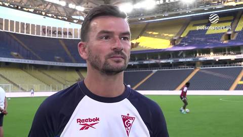 Michal Gasparik, ubrany w str&oacute;j sportowy z logo Reebok i Czuwaj, stoi na tle stadionu piłkarskiego z ż&oacute;łtymi i niebieskimi trybunami. W tle widać murawę, innych piłkarzy i logo Polsat Sport.