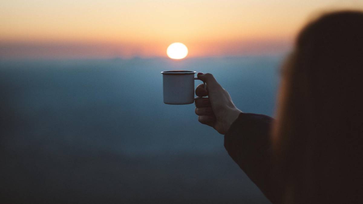 Dłoń trzymająca kubek na tle zachodzącego słońca.