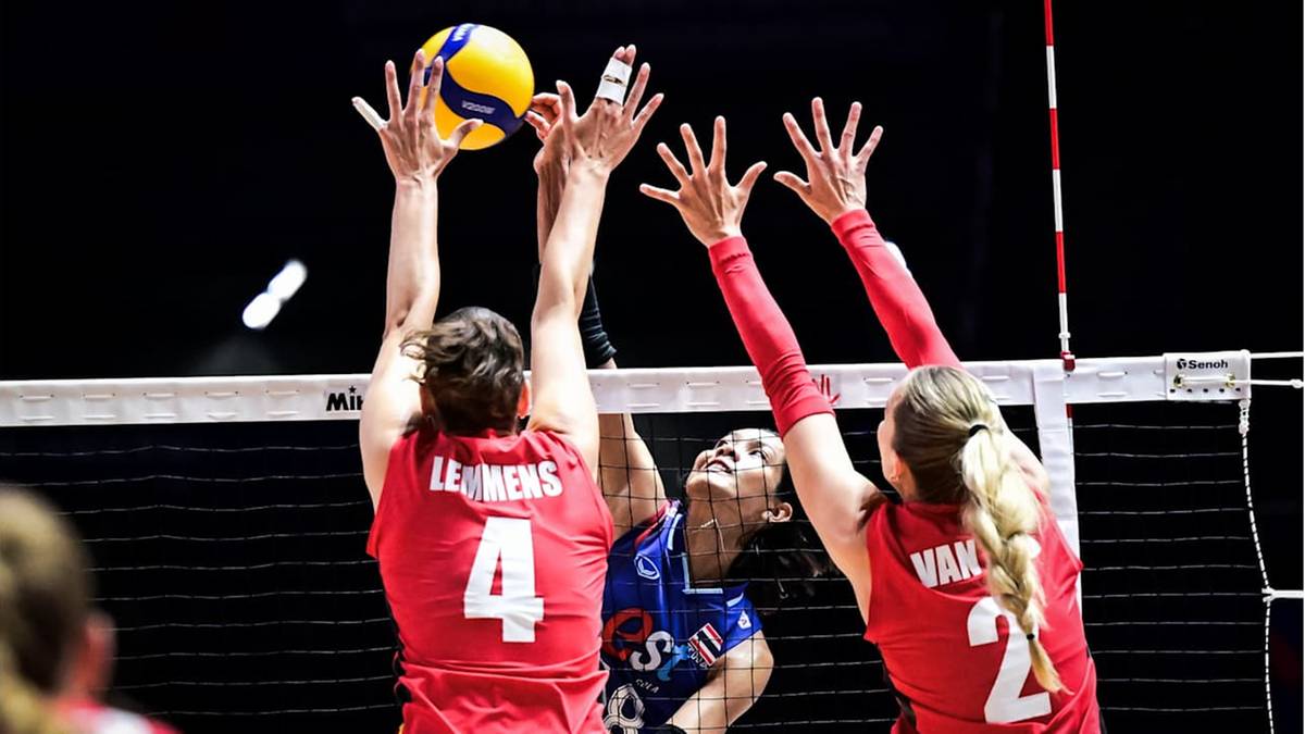 Trzy zawodniczki siatk&oacute;wki skaczą do bloku. Jedna z nich, w czerwonej koszulce z numerem 4, odbija piłkę ręką w kierunku siatki.
