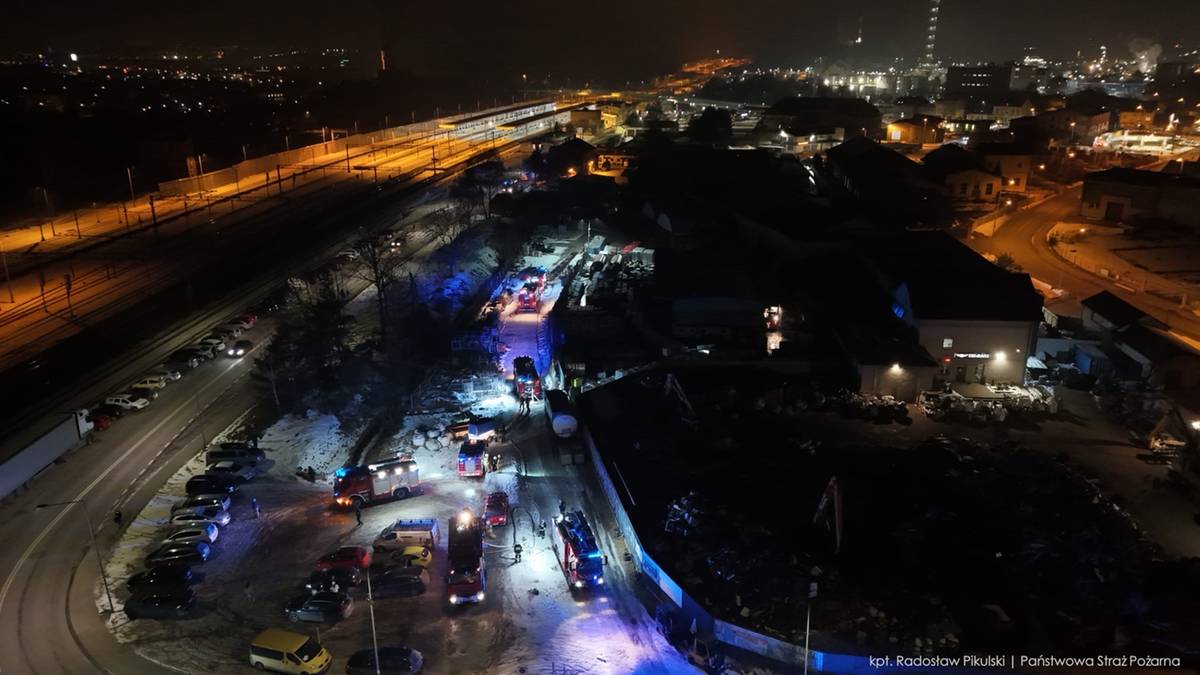 Nocny widok z lotu ptaka na akcję gaśniczą, z widocznymi wozami strażackimi i płonącym budynkiem magazynowym.