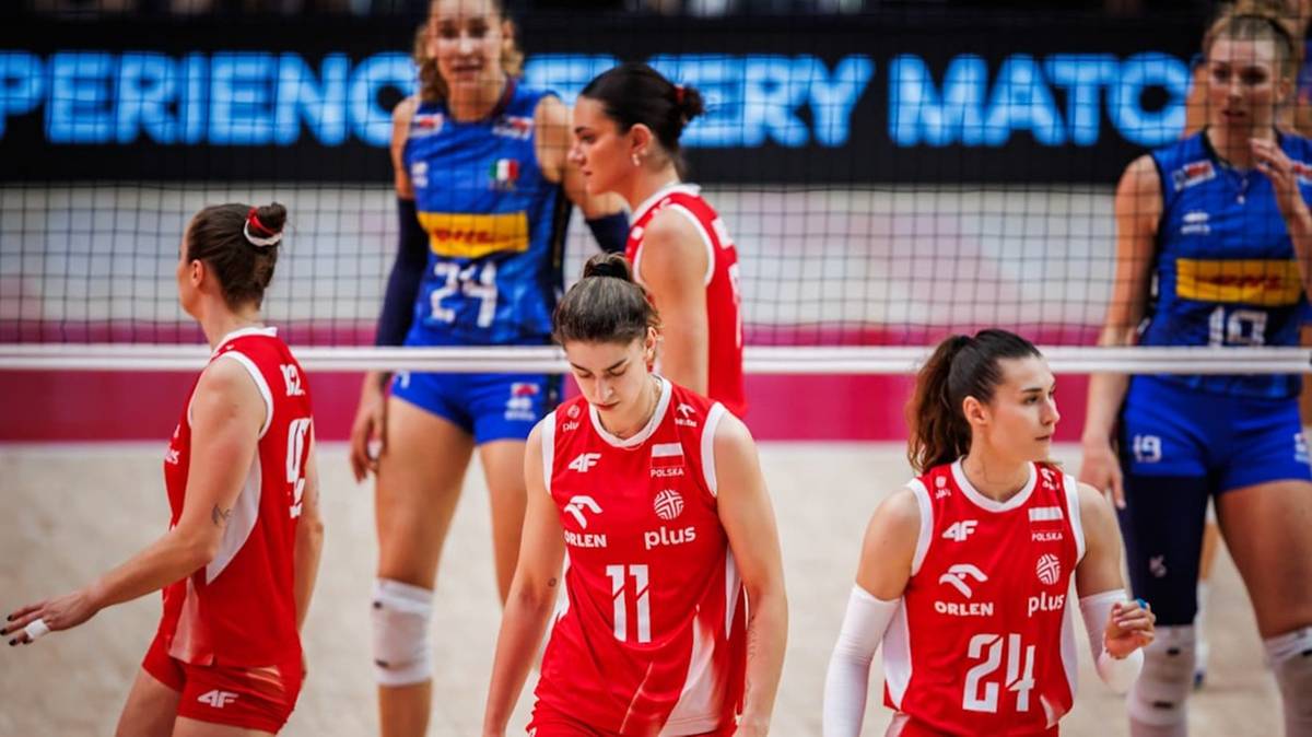 Na zdjęciu grupa siatkarek w czerwonych i niebieskich strojach na tle siatki, niektóre z nich patrzą w dół.