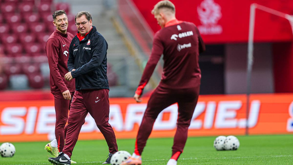 Jan Urban, trener piłkarski, stoi na boisku i rozmawia z dwoma zawodnikami w bordowych strojach treningowych.
