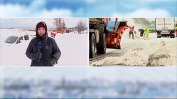 Dziennikarka w zasypanym śniegiem miejscu, obok pogrzebany samochód oraz widok na drogę z pługiem śnieżnym i pracującymi ludźmi.