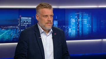 Adrian Zandberg w ciemnej marynarce i białej koszuli na tle niebieskiego ekranu telewizyjnego.